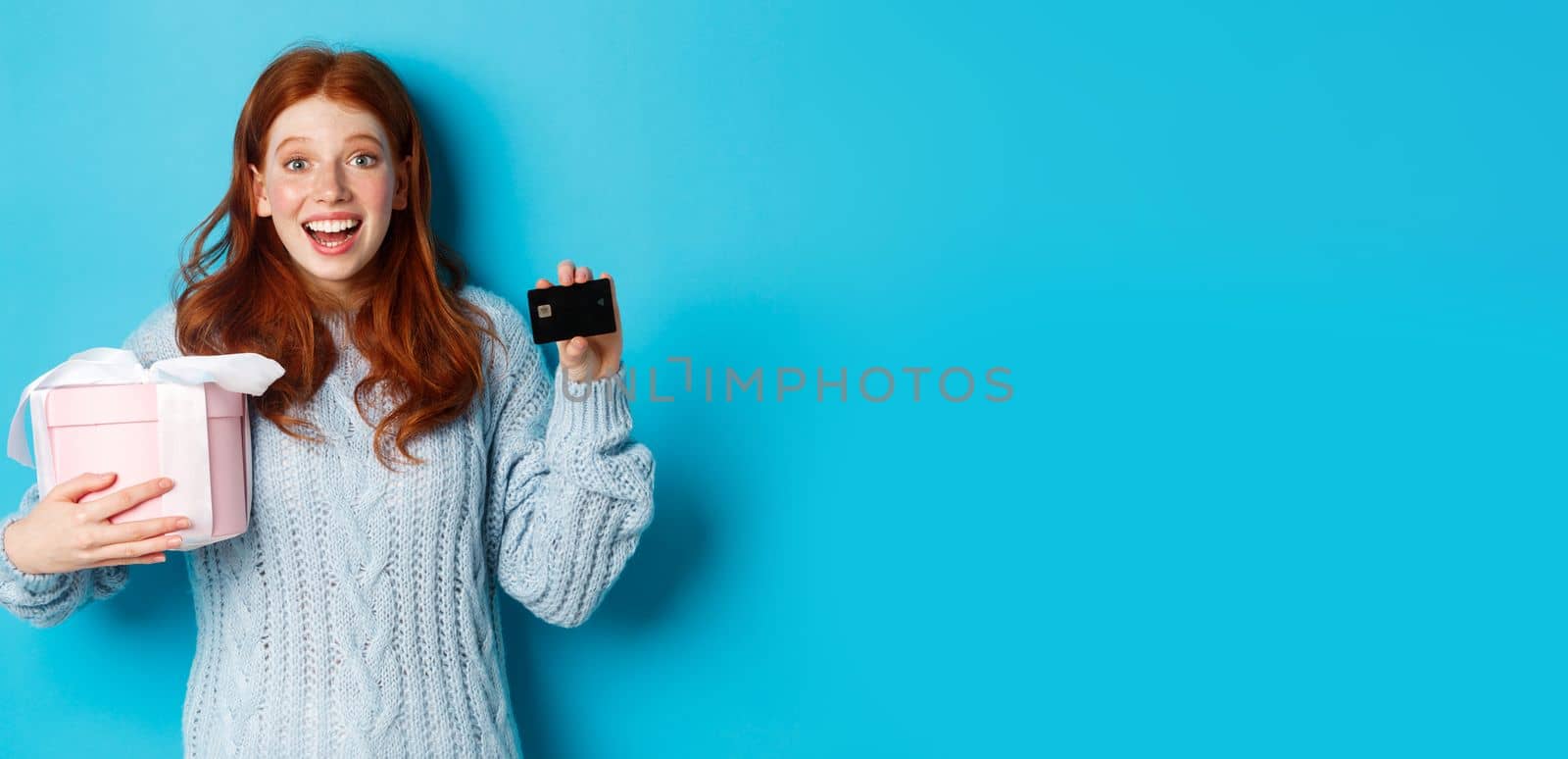 Winter holidays promo offer concept. Cheerful redhead woman holding Christmas gift and credit card, staring at camera amazed, standing over blue background.