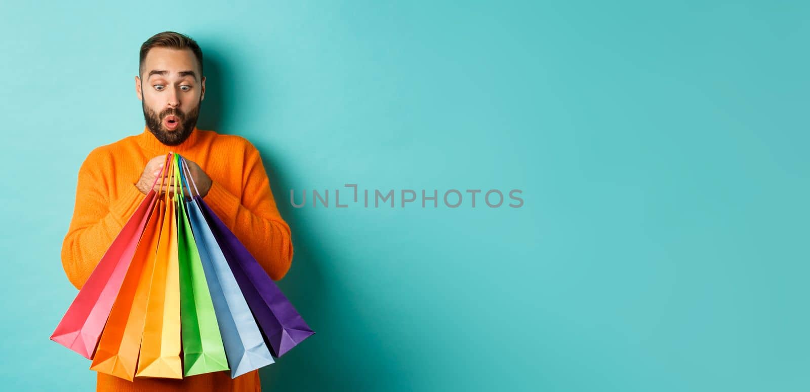 Excited man looking at shopping bags with purchases, standing amused against turquoise background. Copy space