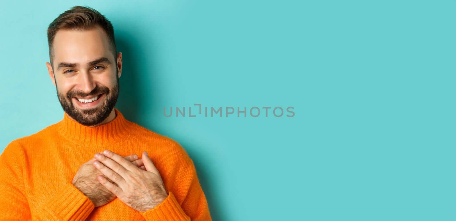 Close-up of handsome young man saying thank you, holding hands on heart and smiling grateful, standing over light blue background by Benzoix