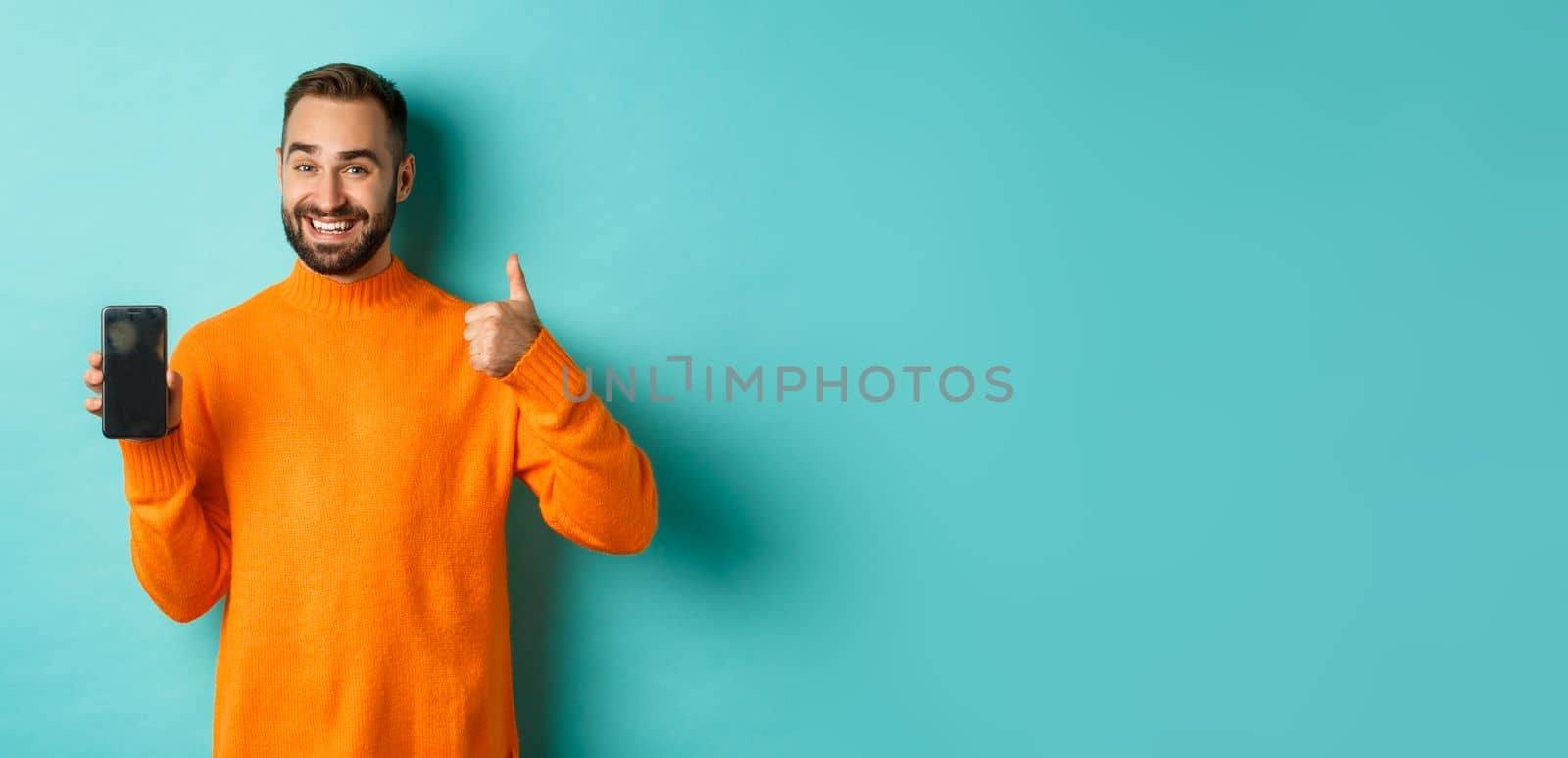 Attractive bearded man showing smartphone screen, thumbs up, recommending mobile app, standing satisfied over turquoise background.