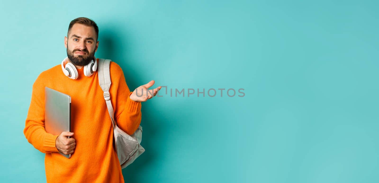 Image of troubled young man with headphones and backpack, shrugging confused and holding laptop, standing over light blue background.