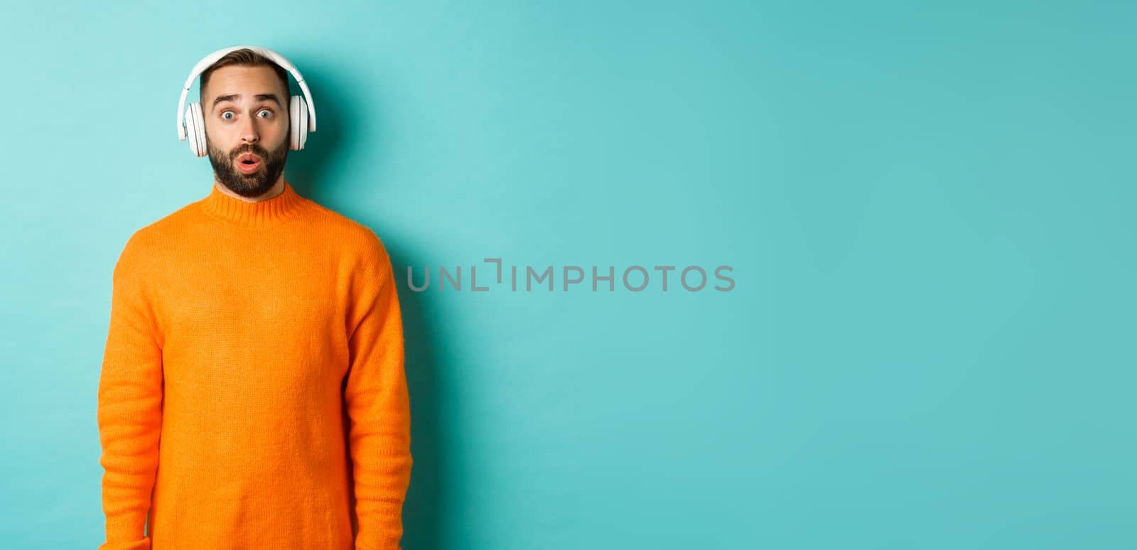 Amazed adult man listening music in headphones, looking at camera impressed with sound, standing over turquoise background.