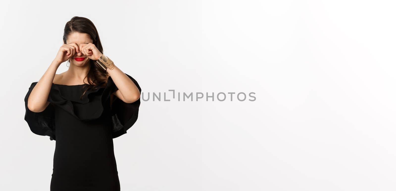 Fashion and beauty. Sad and gloomy woman in black dress crying, rubbing eyes and sulking, standing over white background.