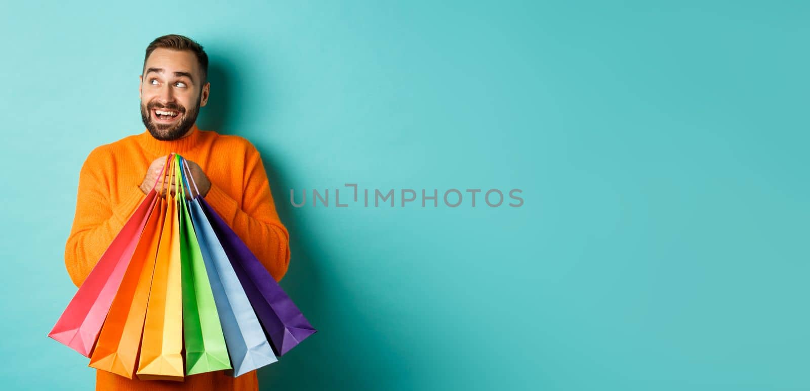 Happy caucasian man in orange sweater, looking left and imaging, holding shopping bags and smiling, standing over turquoise background by Benzoix