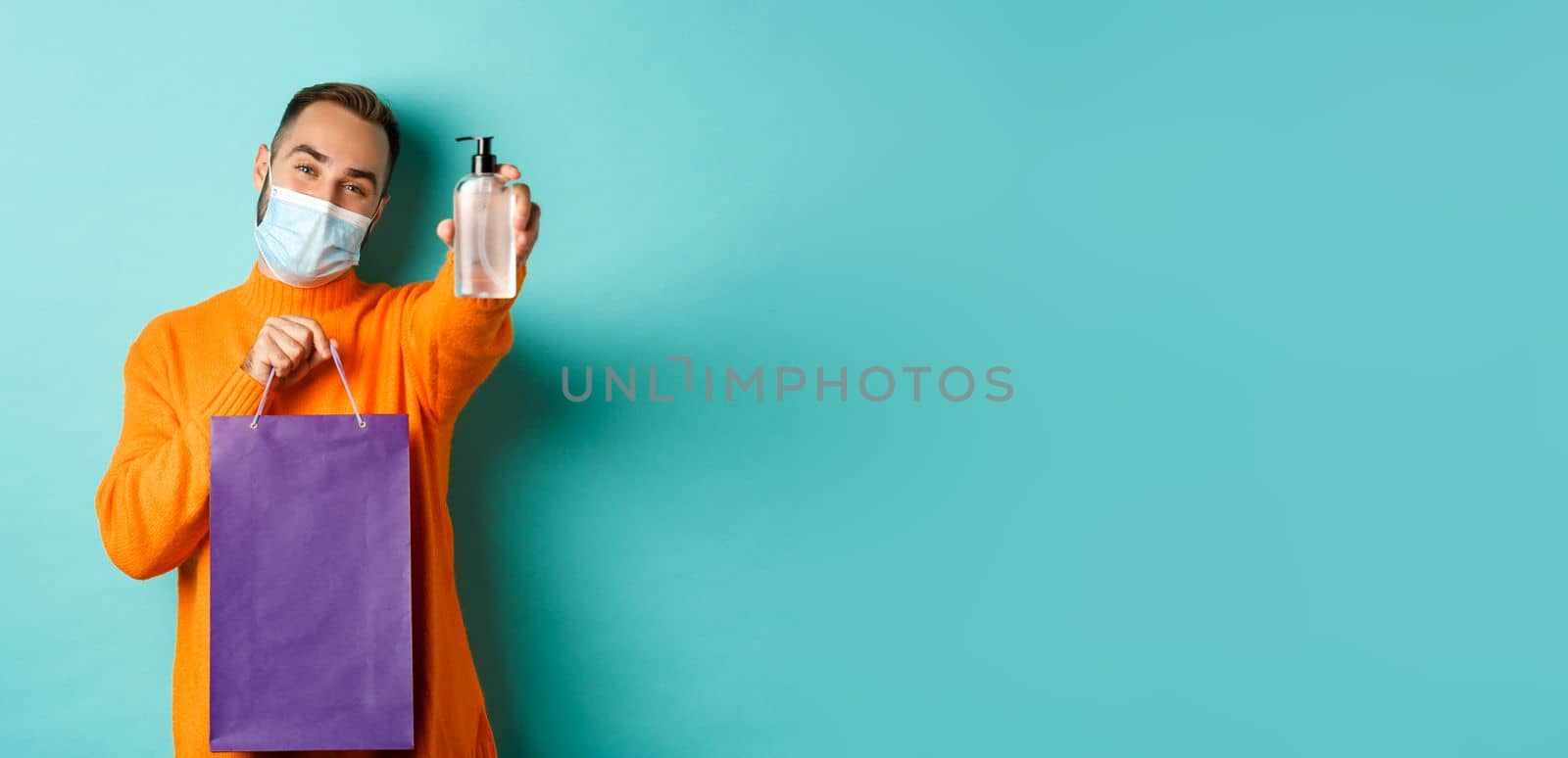 Coronavirus, pandemic and lifestyle concept. Man in face mask showing shopping bag and hand sanitizer, standing over turquoise background.