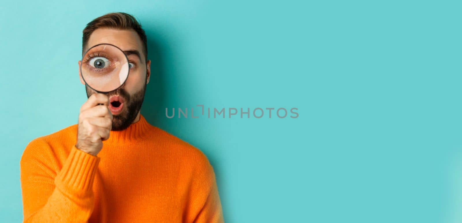 Funny man looking through magnifying glass, searching or investigating something, standing in orange sweater against turquoise background by Benzoix