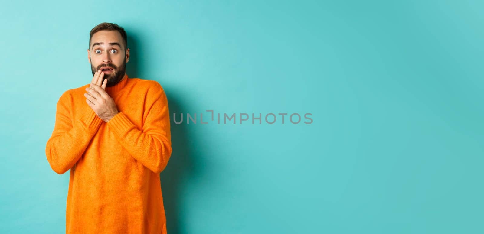 Scared and shocked guy looking at camera, standing in orange sweater against turquoise background by Benzoix