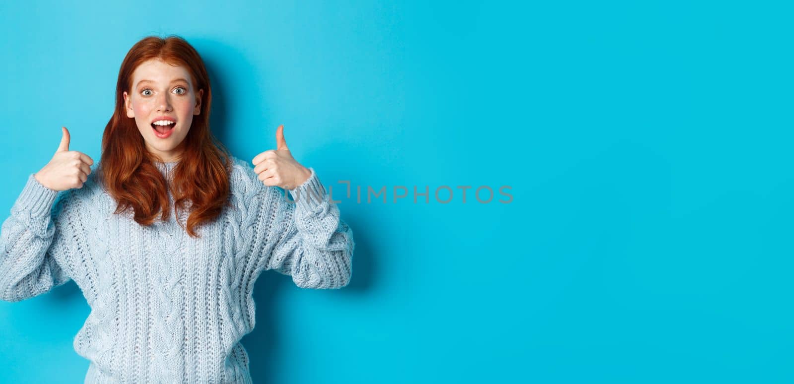 Amazed redhead girl in sweater, showing thumbs up and praising product, looking at camera amazed, standing over blue background. Copy space