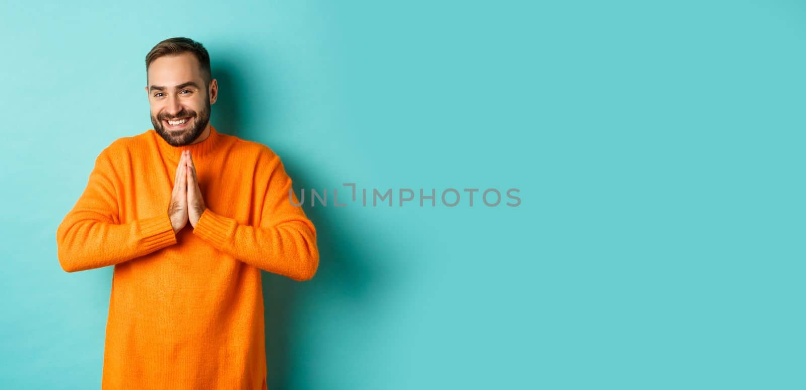 Happy man saying thank you, holding hands in pray, looking grateful and smiling, standing over light blue background.