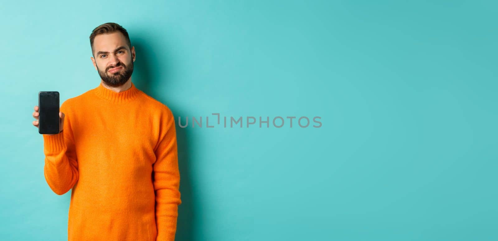 Displeased and disappointed man in orange sweater, grimacing and showing mobile screen, standing over light blue background by Benzoix