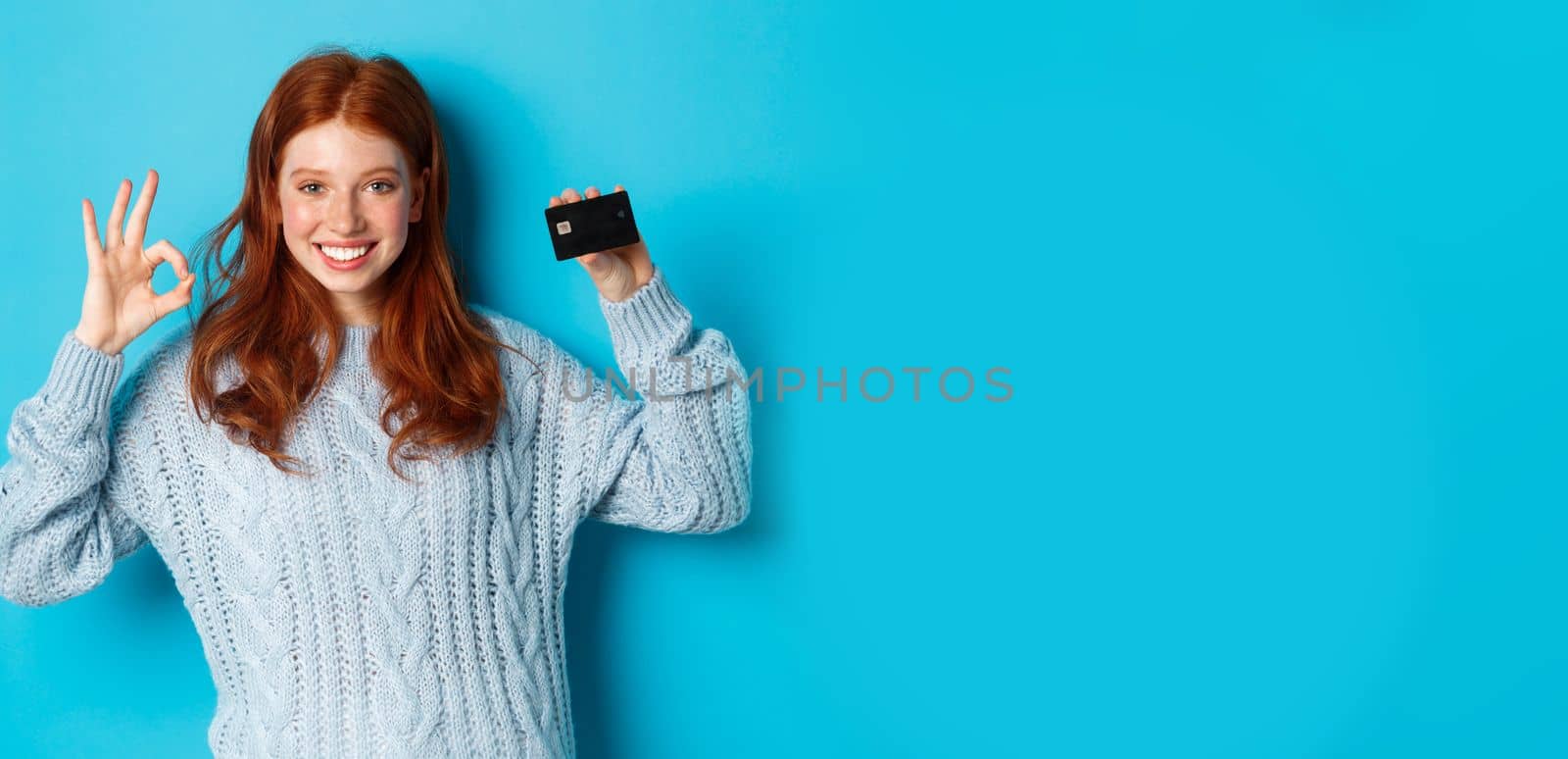 Happy redhead girl in sweater showing credit card and okay sign, recommending bank offer, standing over blue background.