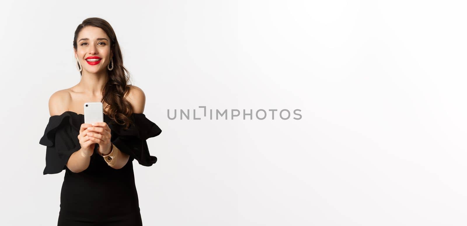 Attractive party girl taking photo on smartphone, make photograph on mobile phone camera, standing in black dress over white background.