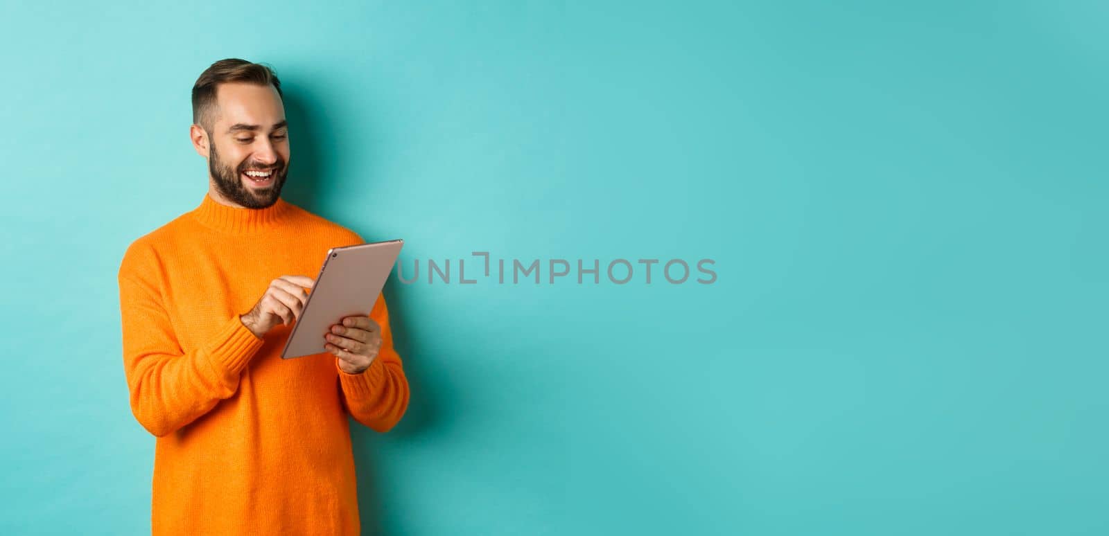 Handsome adult man working on digital tablet and smiling, shopping online, standing over light blue background.