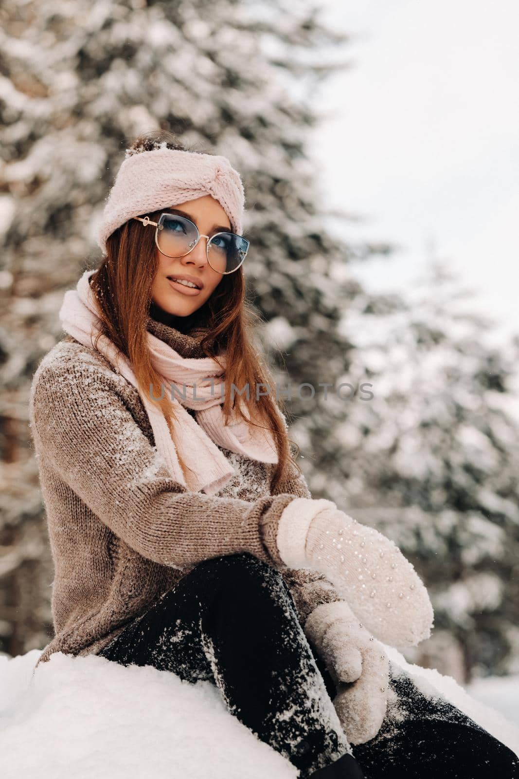 A girl in a sweater and glasses in winter sits on a snow-covered background in the forest by Lobachad