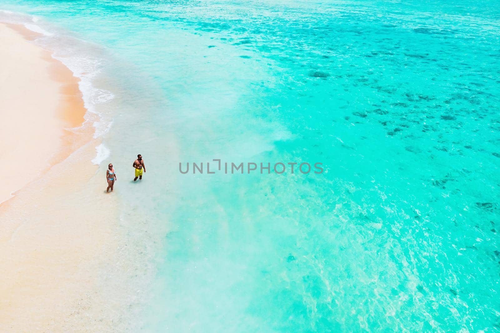 An aerial view of a couple standing on a tropical beach with a view of breaking waves on a tropical Golden sand beach. Sea waves gently loop along the beautiful sandy beach.