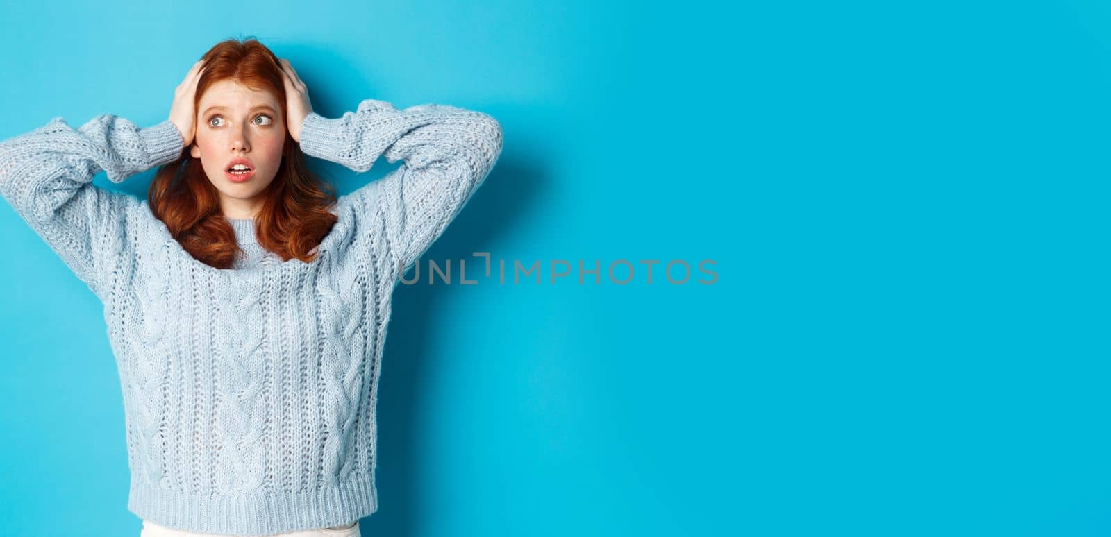 Worried redhead girl standing overwhelmed, holding hands on head in panic and staring left at logo, standing anxious against blue background.