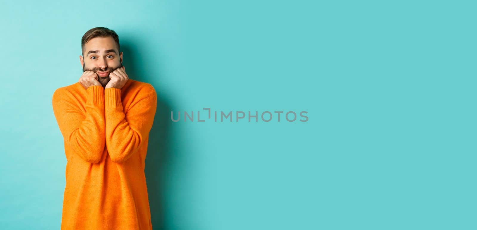 Sad man sighing and looking thoughtful, standing in orange sweater against turquoise background.