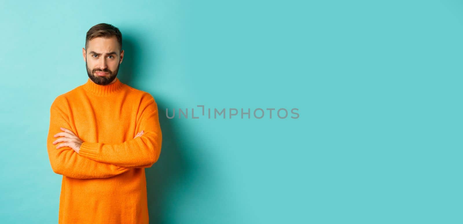 Silly sad guy with beard, sulking and looking offended, cross arms on chest and frowning upset, standing over light blue background.