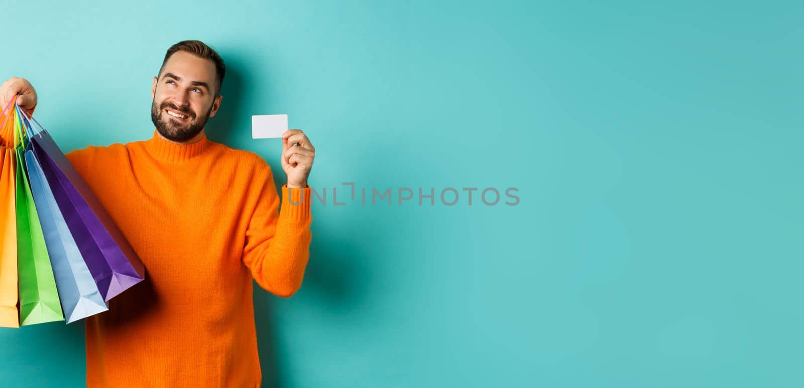 Happy aduly man showing credit card and shopping bags, standing against turquoise background.