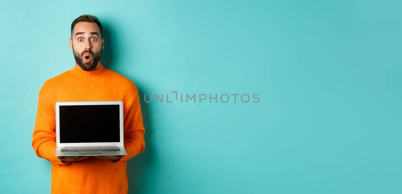 Handsome bearded man in orange sweater showing laptop screen, demonstrating promo, standing over light blue background by Benzoix