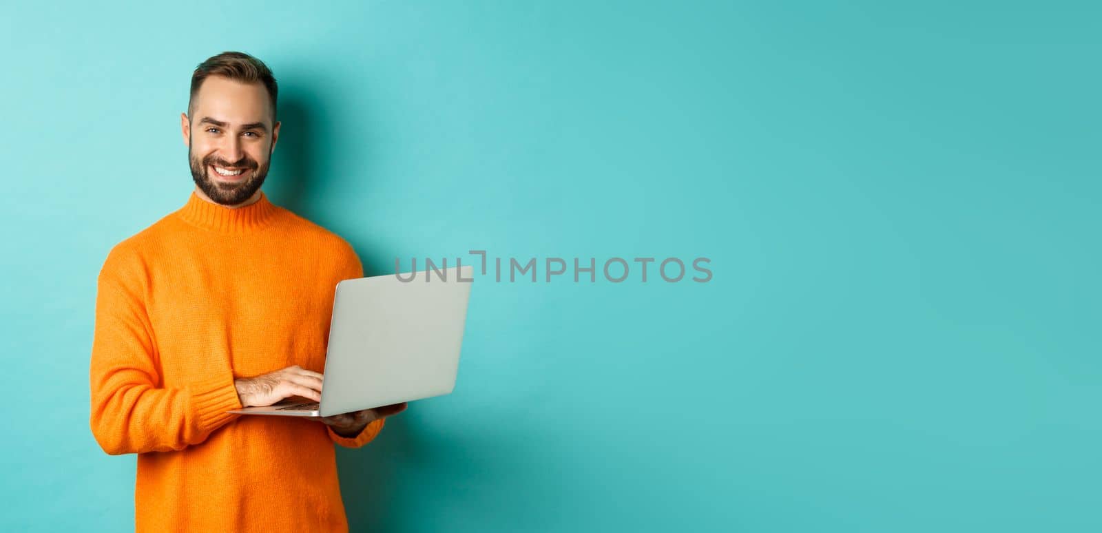 Handsome adult man freelancer working with laptop, smiling at camera, typing on computer keyboard, standing over light blue background by Benzoix