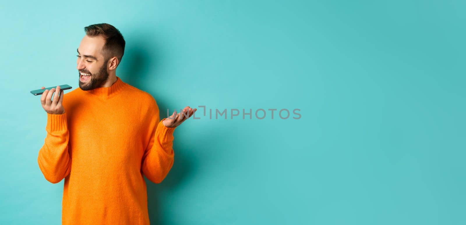 Happy man talking on speakerphone, gesturing and recording voice message on mobile phone, standing in orange sweater over light blue background.