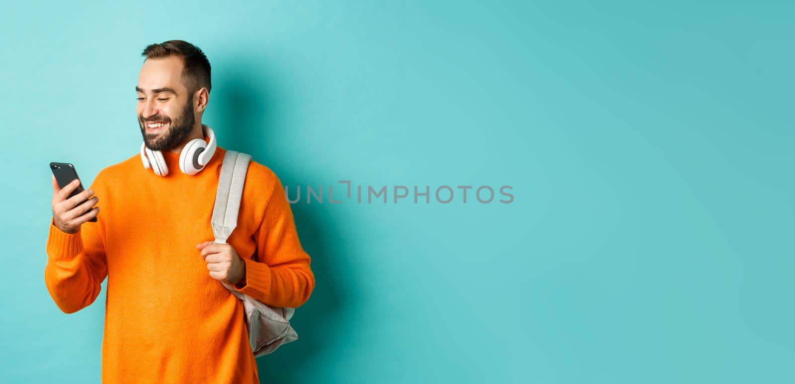 Caucasian man with headphones and backpack looking at phone, reading message and smiling, standing over turquoise background.