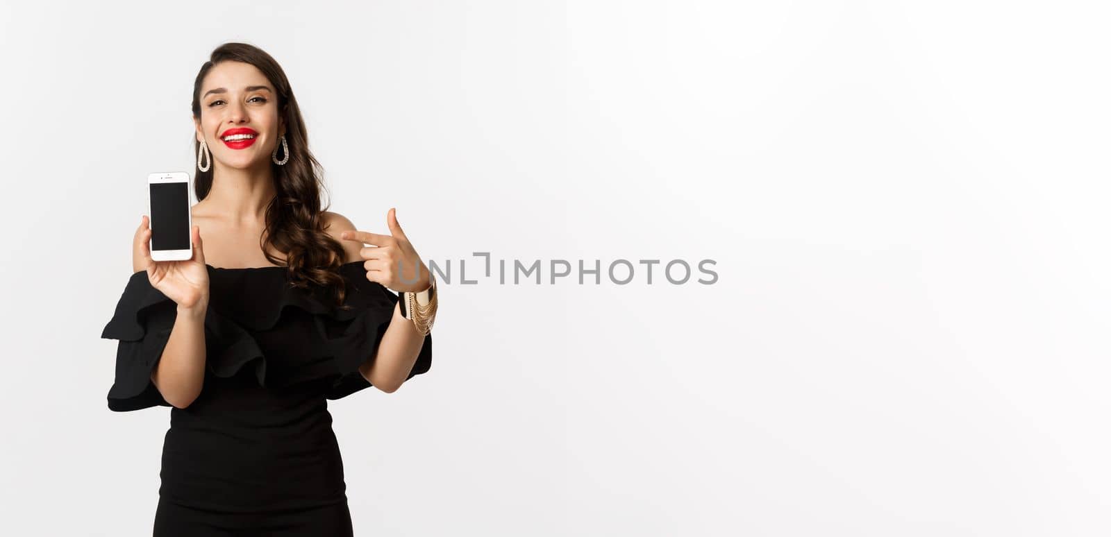 Online shopping concept. Fashionable woman in black dress pointing finger at smartphone screen, showing application, standing over white background.