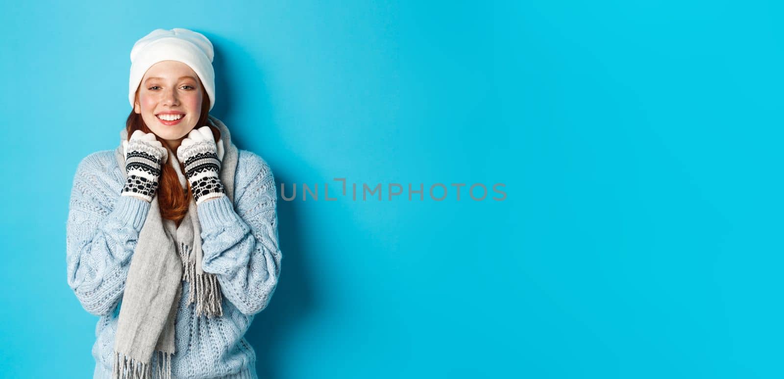Winter and holidays concept. Cute redhead girl in white beanie and gloves smiling at camera, looking delighted, standing against blue background by Benzoix