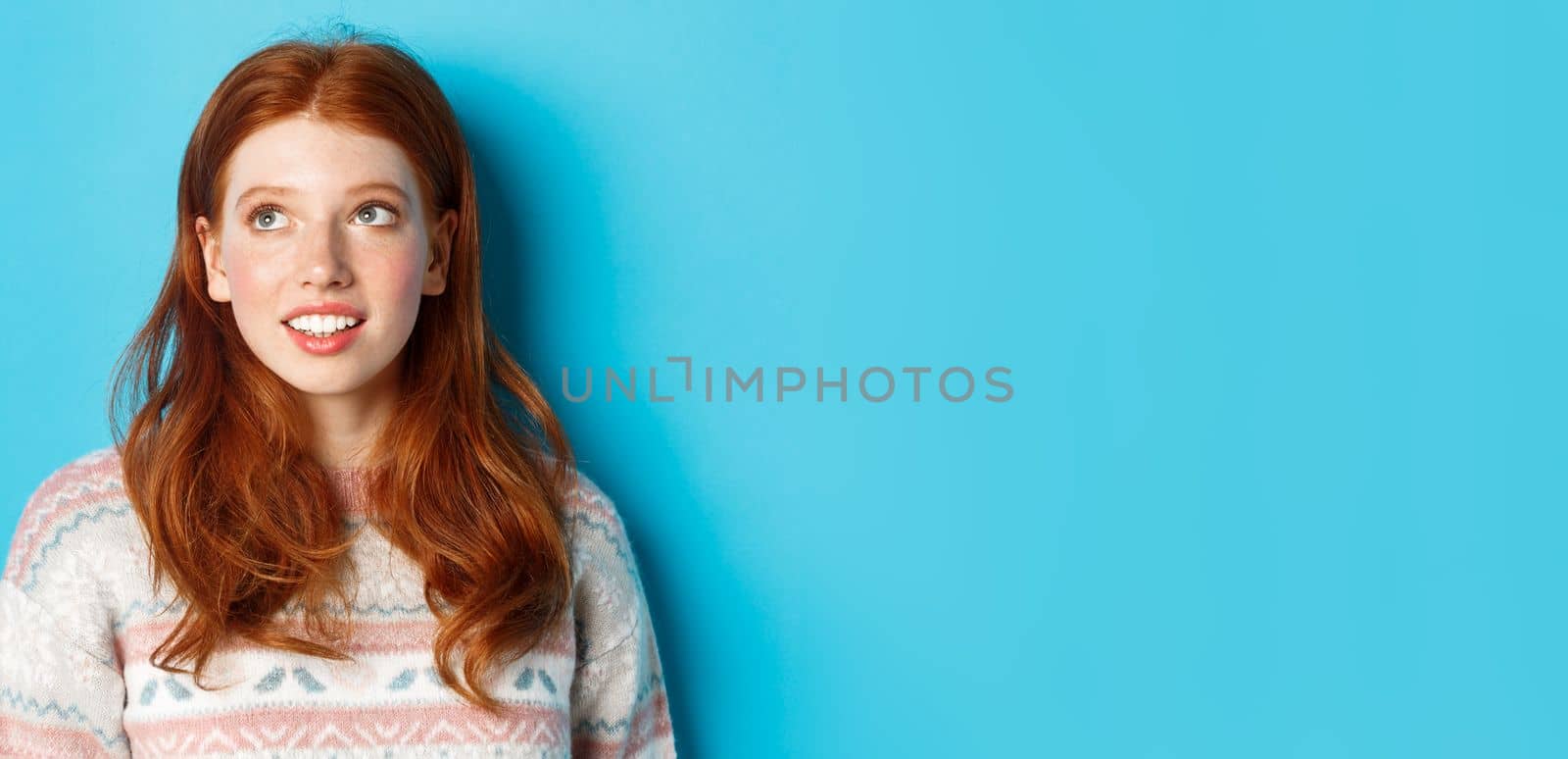 Close-up of dreamy teen girl with red hair, looking at upper left corner and smiling, standing against blue background.