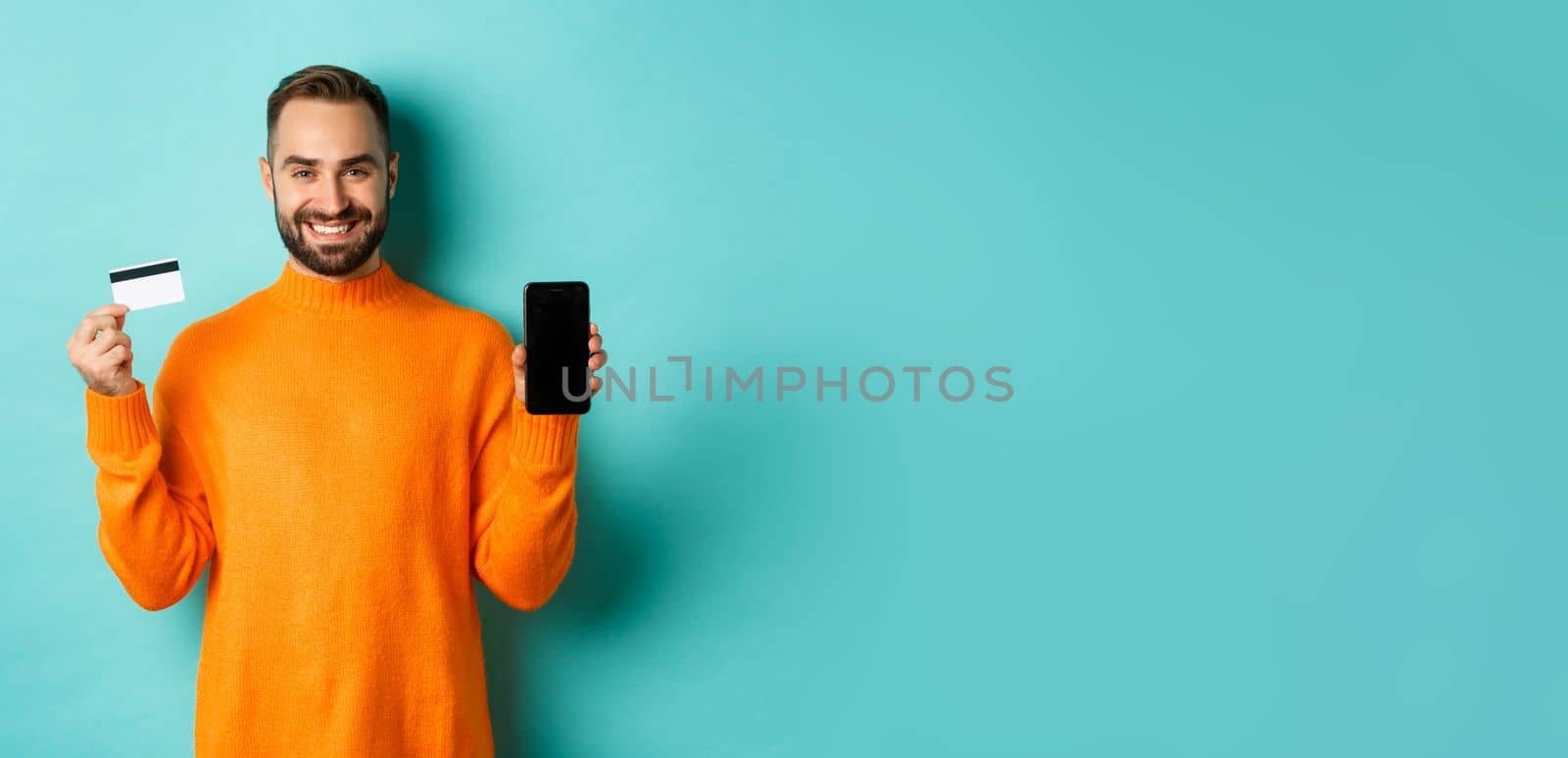 Online shopping. Happy attractive guy showing mobile phone screen and credit card, smiling satisfied, standing over light blue background by Benzoix