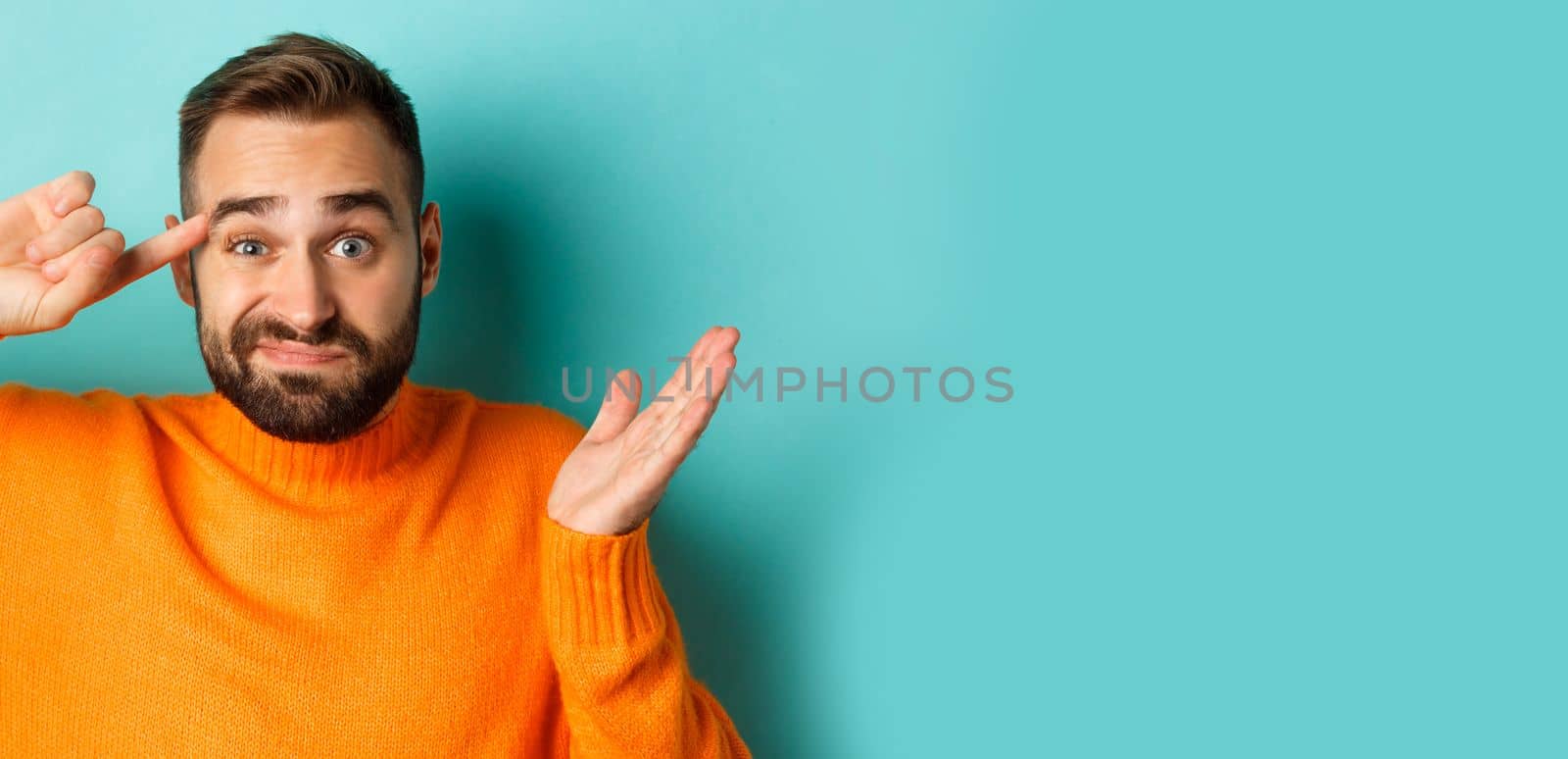 Close-up of man expressing disdain, scolding person, pointing finger at head and looking at camera, standing over light blue background.