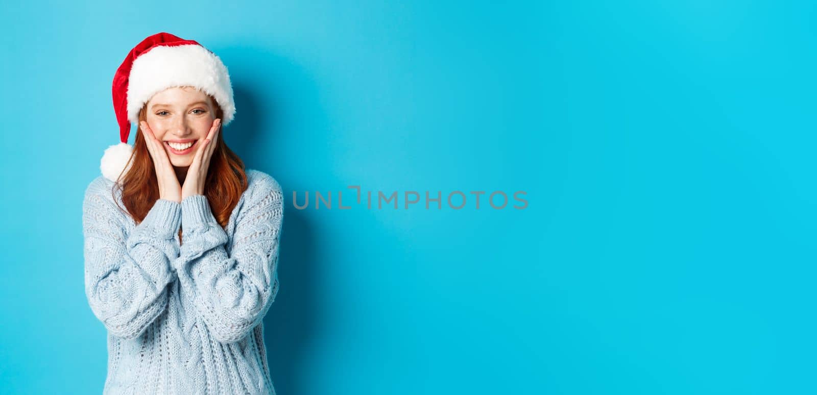 Winter holidays and Christmas Eve concept. Cheerful redhead girl in santa hat, celebrating New Year, blushing and smiling happy, standing over blue background by Benzoix