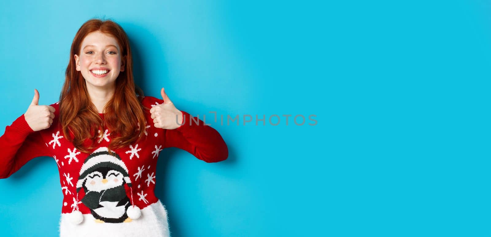Winter holidays and Christmas Eve concept. Happy smiling girl with red hair, showing thumbs up in approval, praise good product, recommending, standing in xmas sweater.