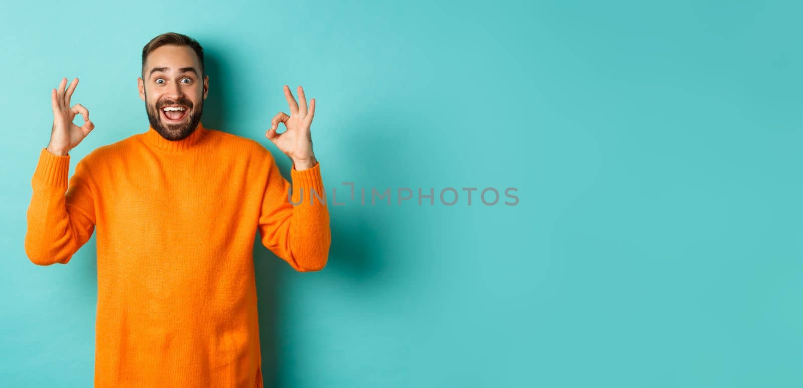 Impressed man praising something awesome, showing okay signs and looking amazed, standing in orange sweater at light blue background by Benzoix