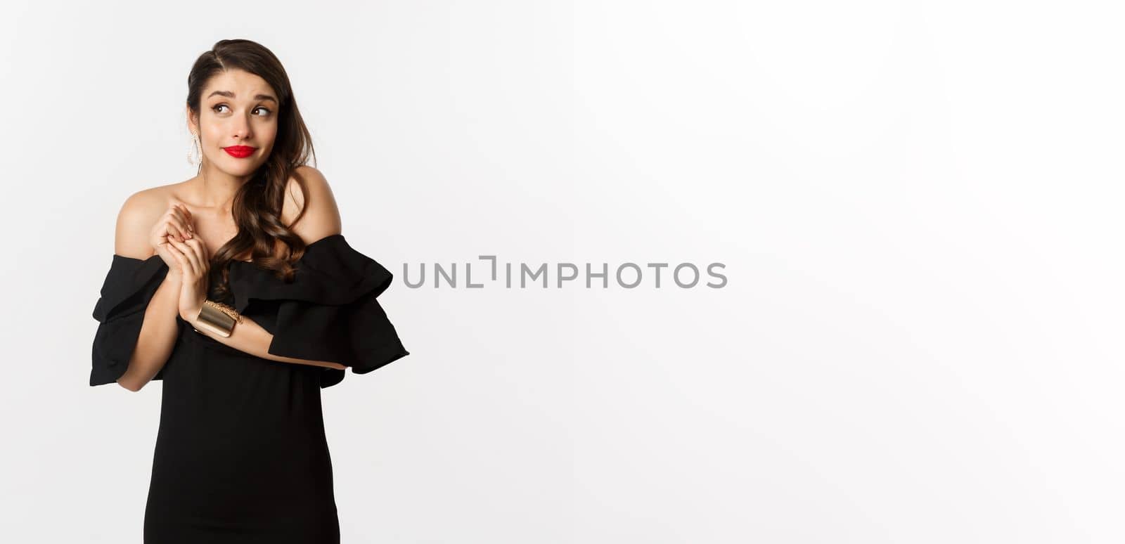Fashion and beauty. Young silly woman in black dress pouting and looking timid, standing in black glamour dress over white background.