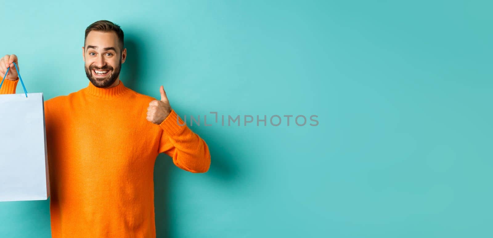 Handsome smiling man showing thumbs-up and shopping bag, recommending store, standing over blue background by Benzoix