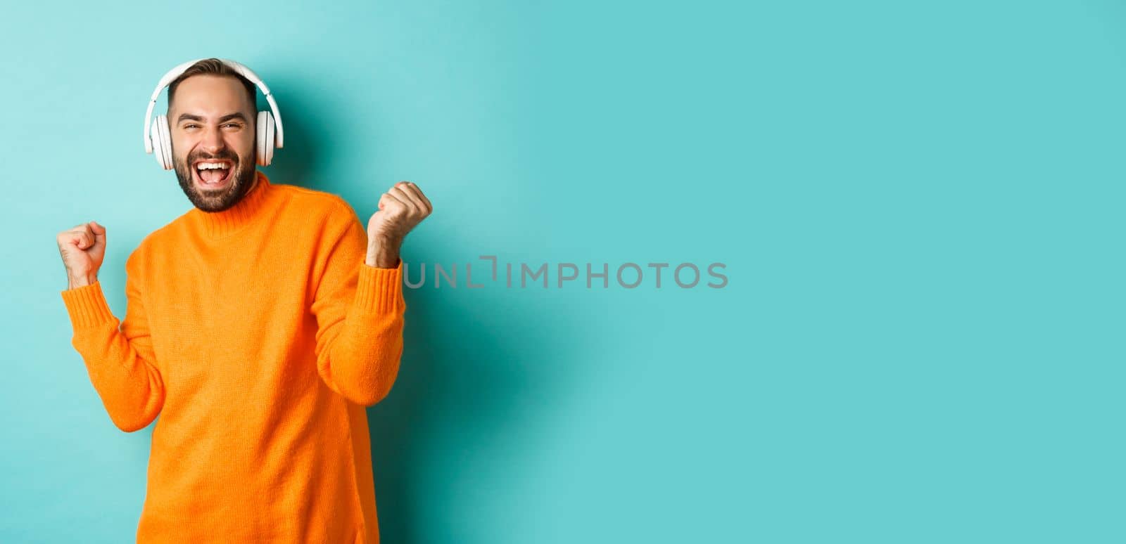 Happy adult man in orange sweater, looking up and listening music in headphones, standing over blue background by Benzoix