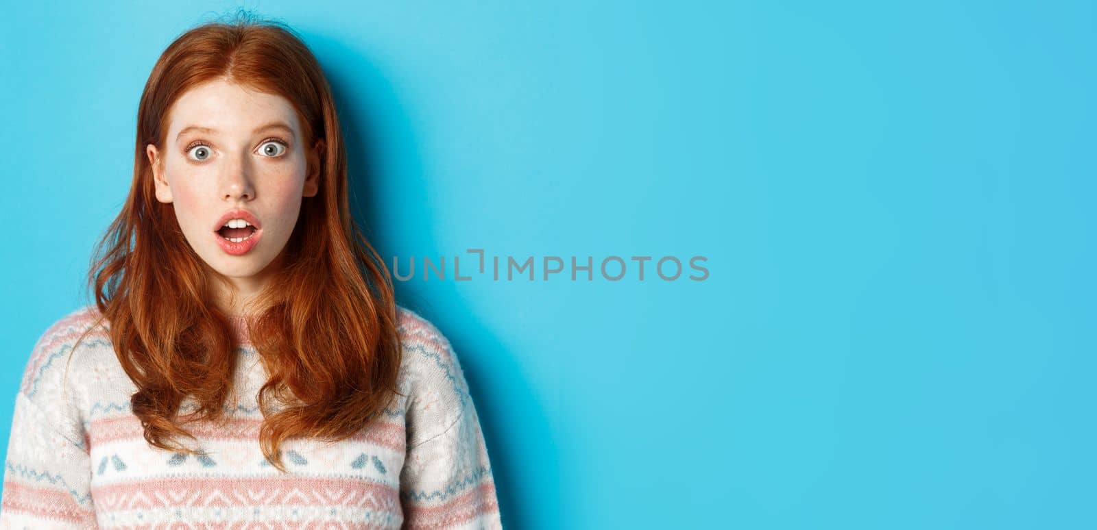 Close-up of shocked redhead girl drop jaw in awe, staring with amazement at camera, standing against blue background by Benzoix