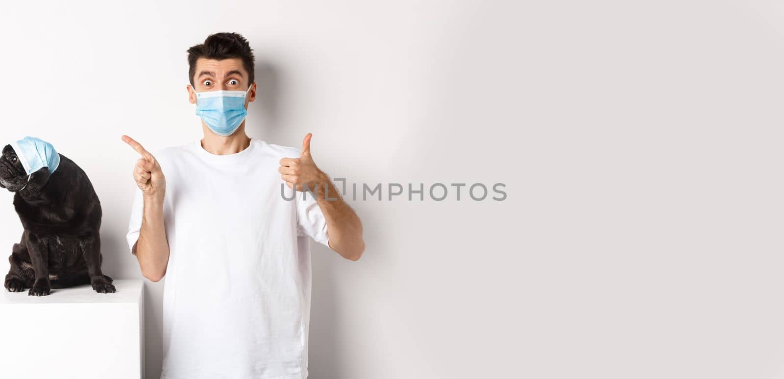 Covid-19, animals and quarantine concept. Young man and black dog wearing medical masks, pug looking at upper left corner and owner showing thumb-up to praise promo by Benzoix