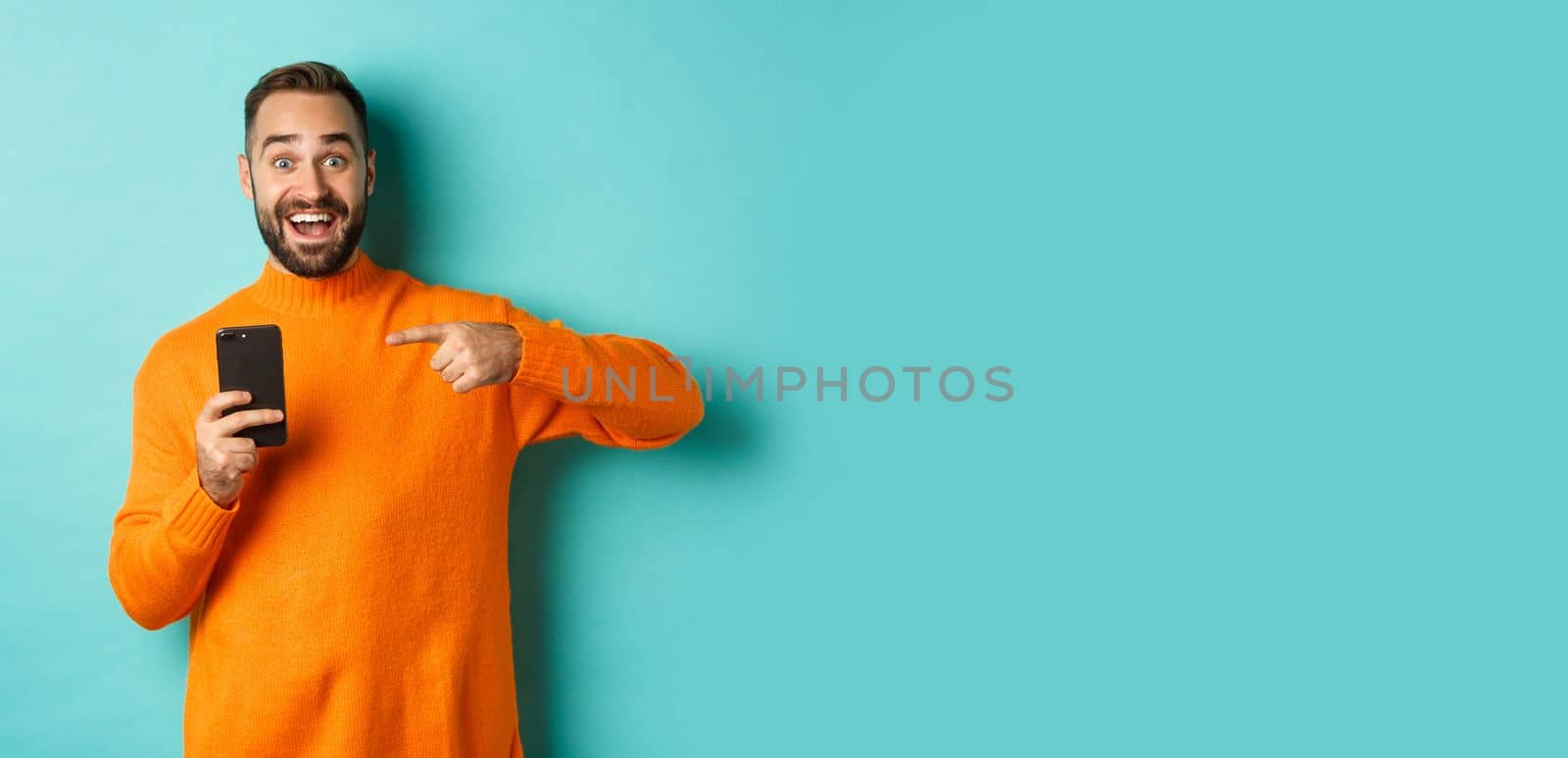 Excited man pointing at mobile phone, showing internet promo, standing over turquoise background, concept of online shopping by Benzoix
