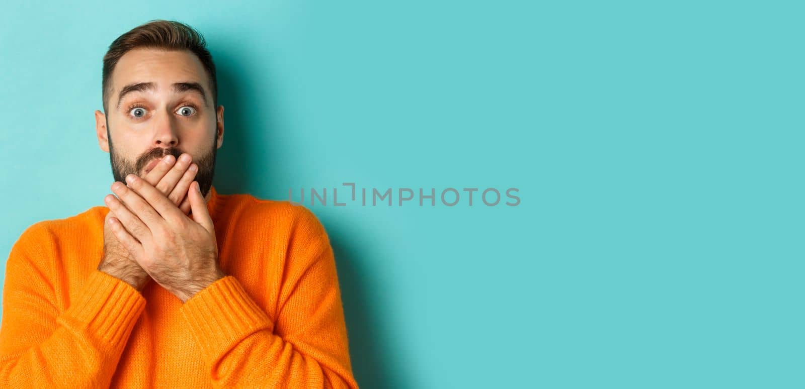 Close-up of shocked caucasian man covering his lips and mouth, staring astounded at camera, standing against turquoise background.