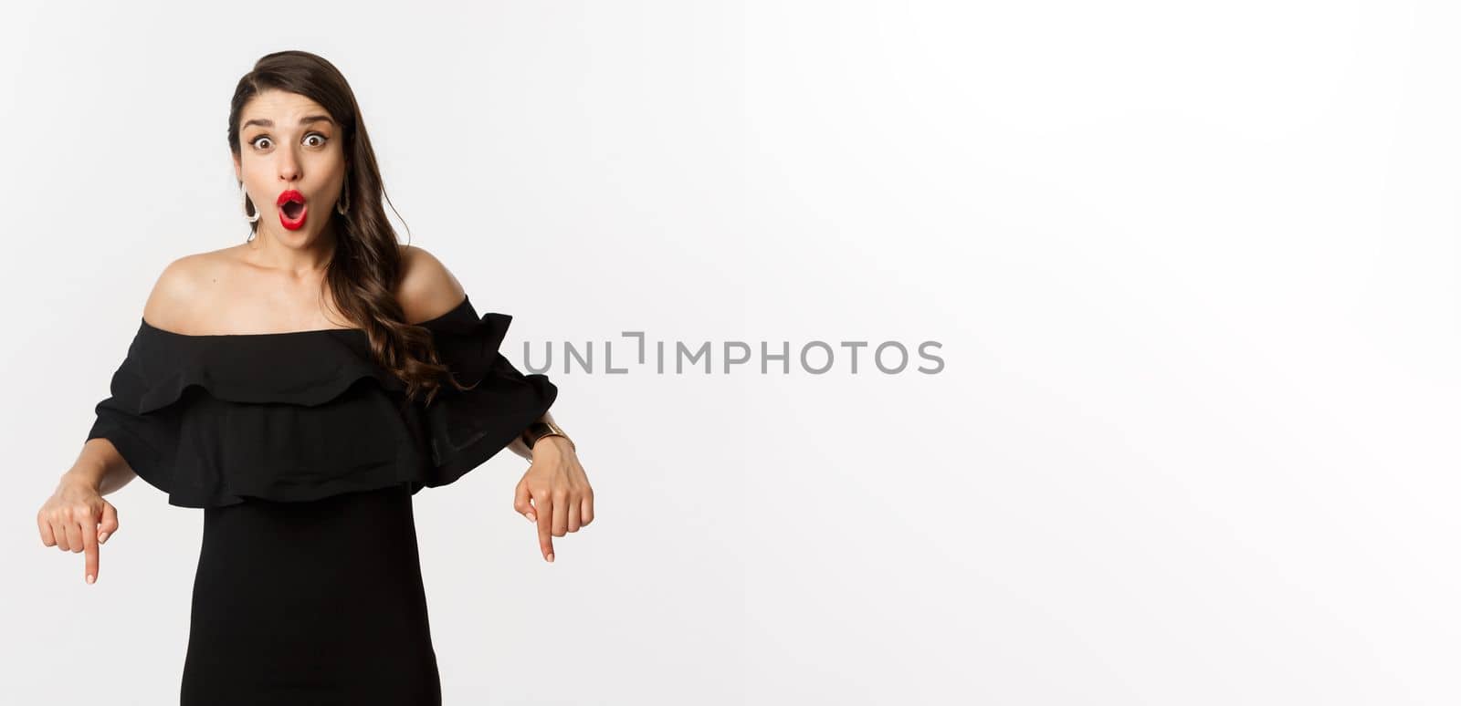 Fashion and beauty. Elegant woman in black dress pointing fingers down, showing promo and smiling, standing over white background.