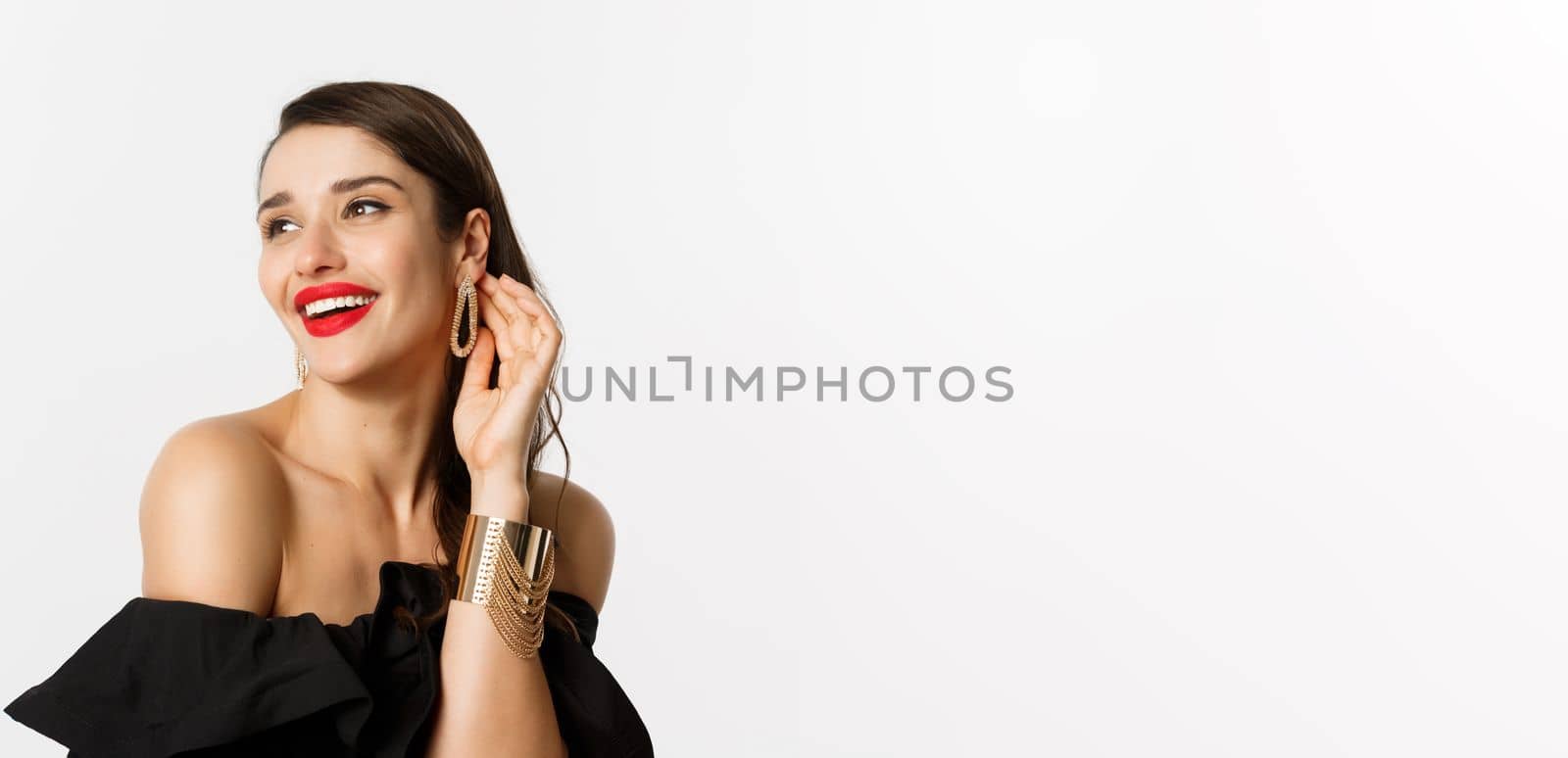 Fashion and beauty concept. Close-up of elegant brunette woman with red lips, black dress, laughing coquettish and gazing away, standing over white background.