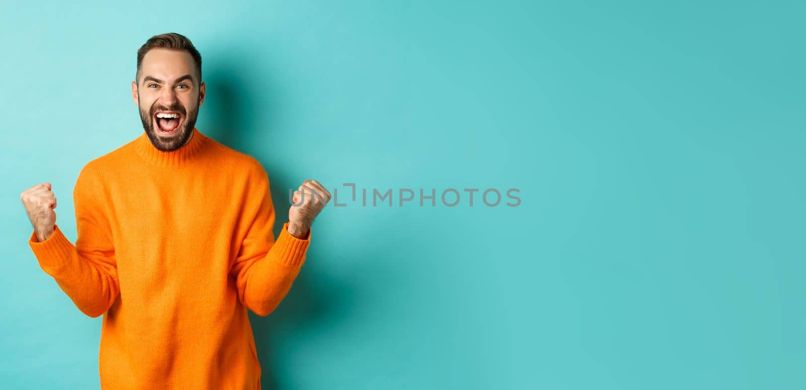 Excited man celebrating victory, rejoicing and making fist pump gesture, winning and looking satisfied, saying yes, achieve goal, standing over light blue background by Benzoix