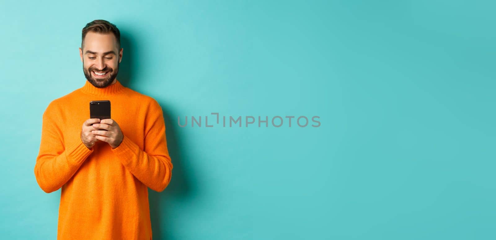 Handsome man smiling and texting message on mobile phone, communicating online, standing over light blue background by Benzoix