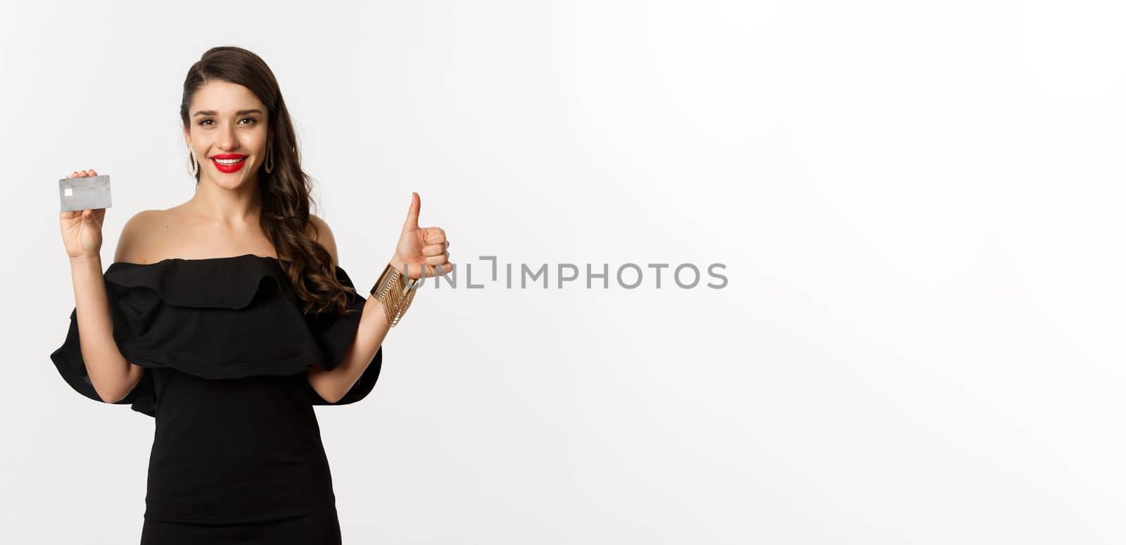 Fashion and shopping concept. Satisfied stylish woman in black dress showing credit card, make thumb-up in approval, recommending, standing over white background.