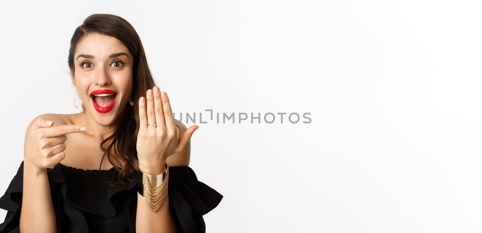 Happy young woman saying yes, become a bride, pointing at engagement ring on finger and smiling pleased, standing over white background.