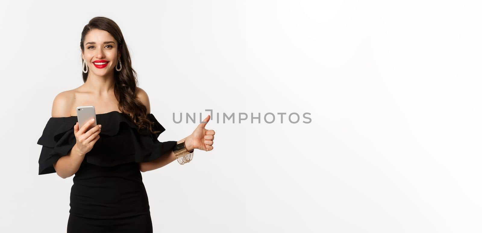 Online shopping concept. Attractive woman in trendy black dress, makeup, showing thumb-up and using mobile phone app, white background.