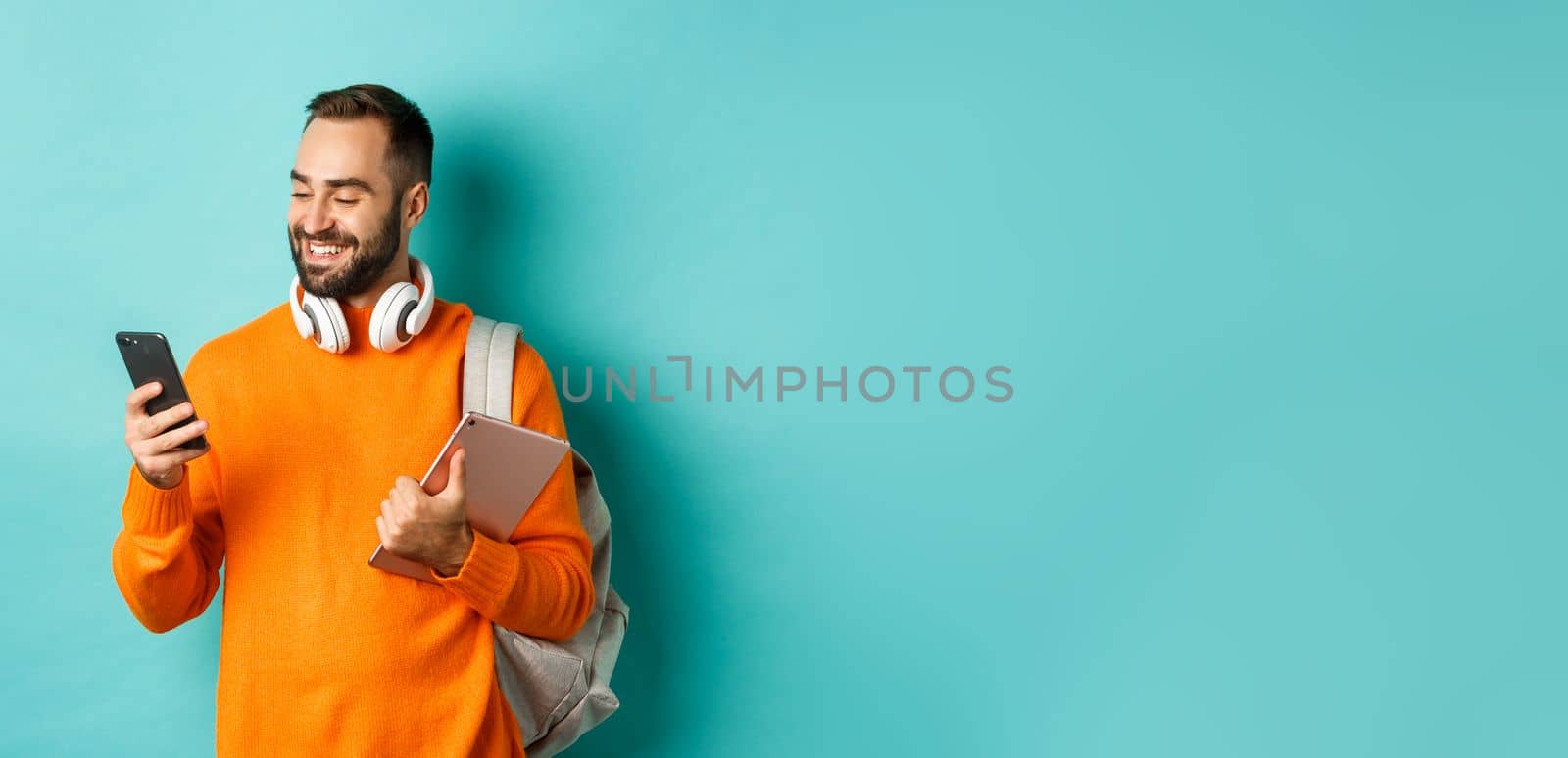 Handsome man student with headphones and backpack, holding digital tablet, reading message on mobile phone, standing against light blue background by Benzoix